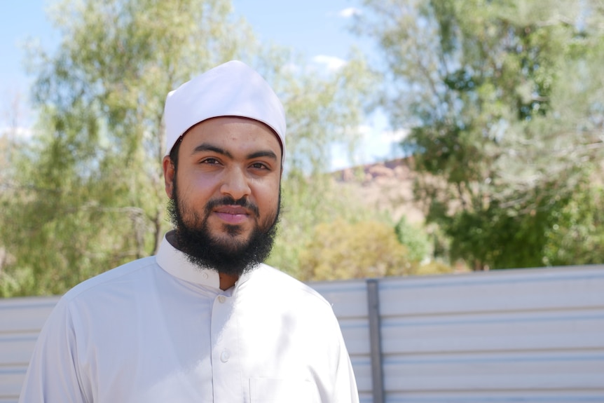 An Imam wearing traditional dress looks at the camera with the red rocky ranges of Alice Springs behind him.