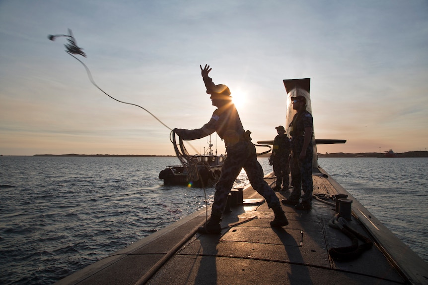 Two men stand on top of the submarine and throw a heaving line to a tug boat.