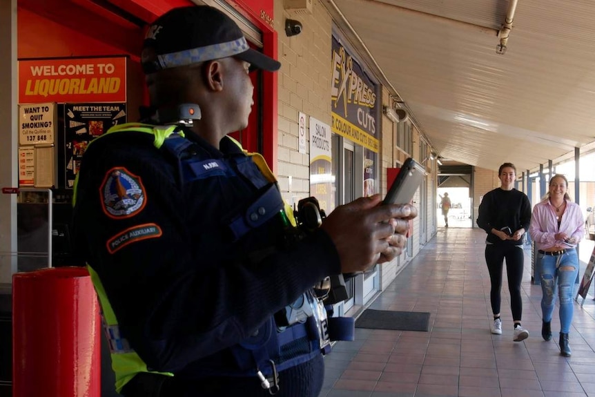 A police auxiliary outside a bottle shop waits for patrons