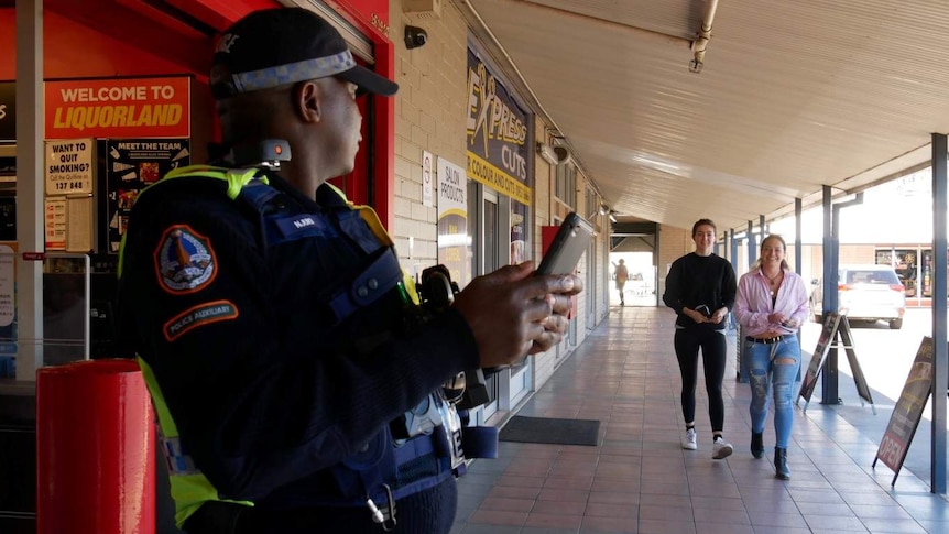 A police auxiliary outside a bottle shop waits for patrons