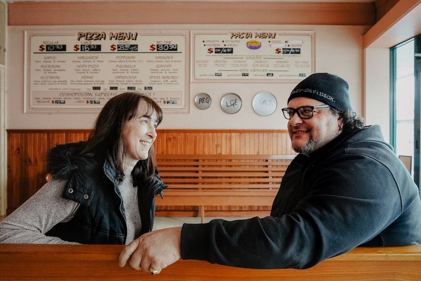A man and a woman sit on a bench in a restaurant smiling at each other, a pizza menu is behind them.