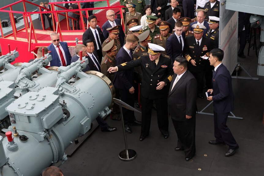 Kim Jong Un stands looking at a frigate surrounded by men in uniform.