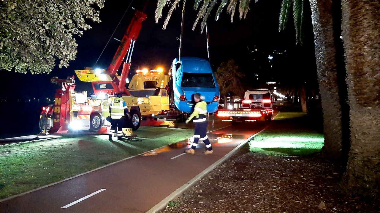 A blue car hangs on a lifting platform next to a crane and recovery vehicle.