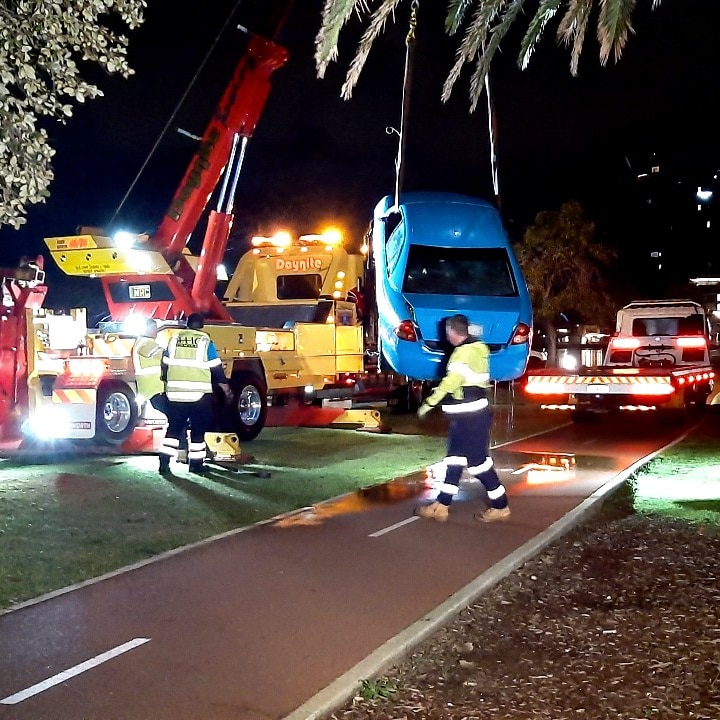 A blue car hangs suspended by a hoist next to a crane and recovery vehicle.