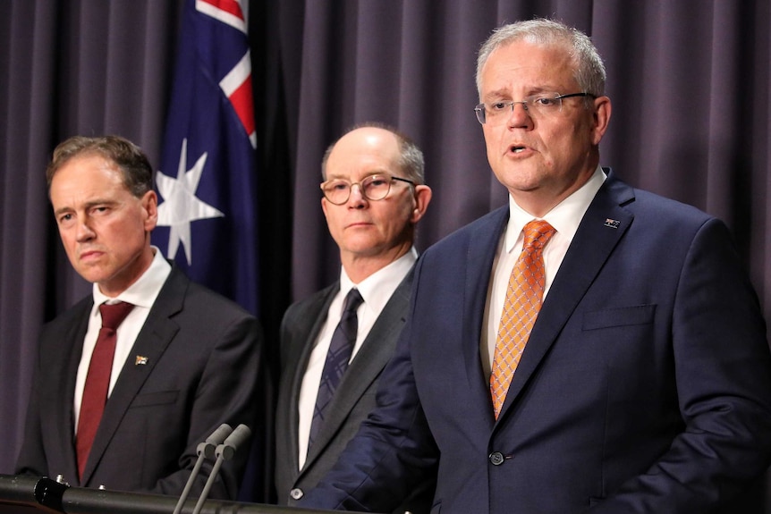 Scott Morrison stands on the podium in the blue room, and Paul Kelly and Greg Hunt speak to the media beside him.