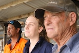A young woman in a blue workshirt looks with concern off camera, standing alongside two older men in workshirts who also look w