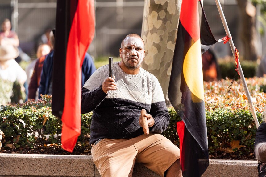 A man sits between two Aboriginal flags, wearing white face paint, banging two wooden sticks — traditional instruments.