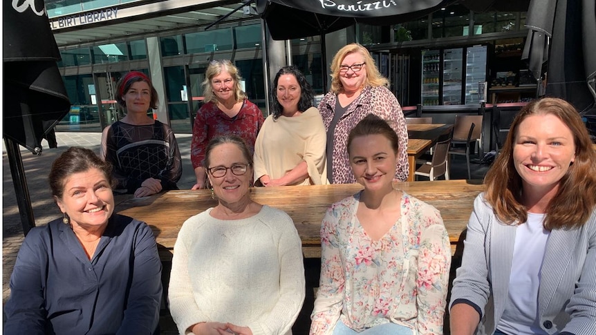 Eight women sitting and standing around a bench outside