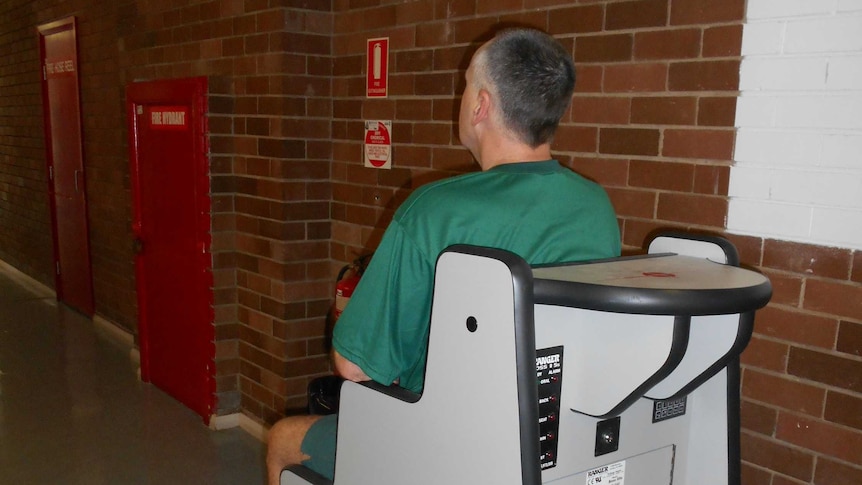 A man in prison greens sits on a body orifice scanner.