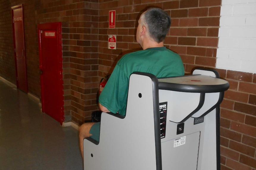 A man in prison greens sits on a body orifice scanner.