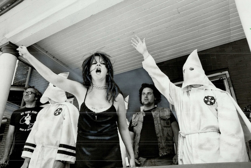 Members of the Ku Klux Klan; on the verandah of a house, raise arms in Nazi-style salute.