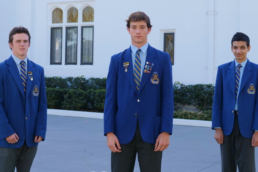 An image of three male students in blue blazers standing in front of a white building