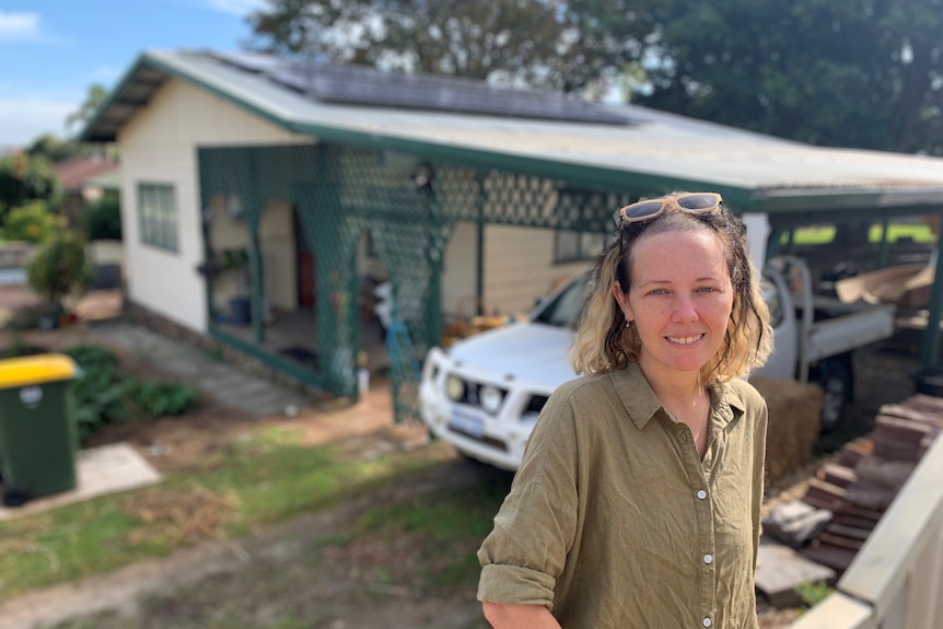 A young woman wearing khaki linen shirt sitting on a fence in front of house with rooftop solar panels.