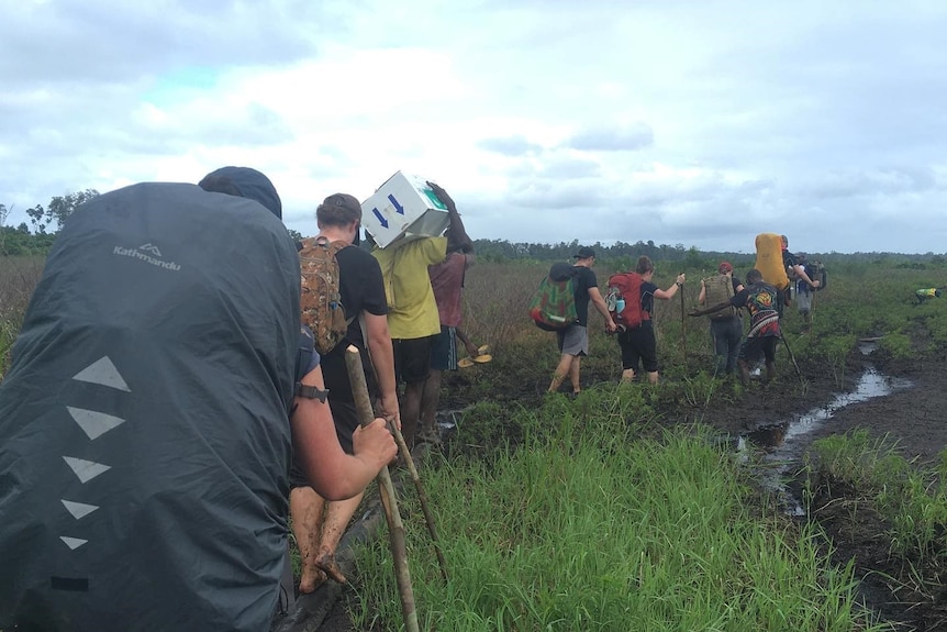 A line of people with large backpacks, walking through grass and mud, using long sticks for stability