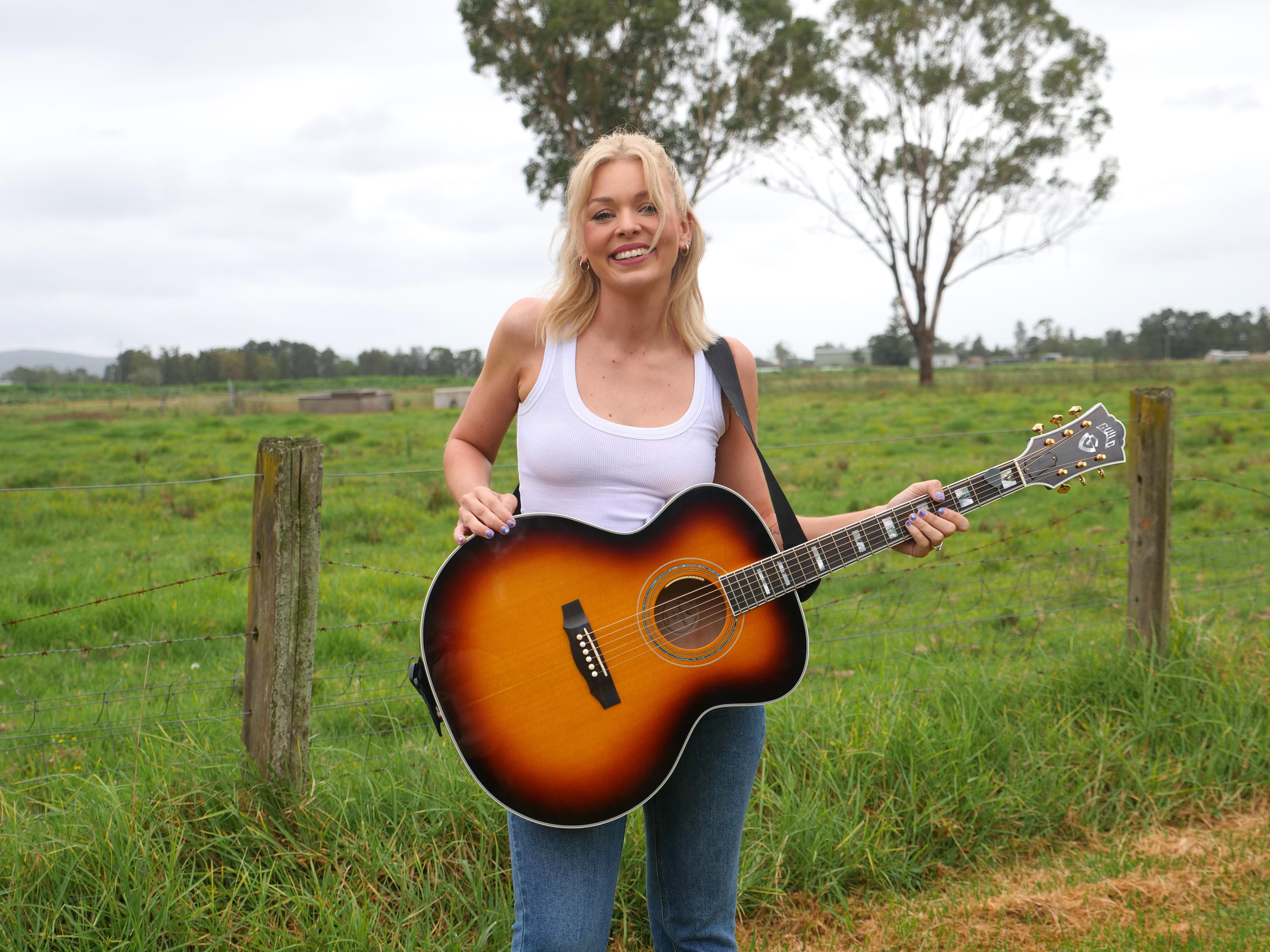 A woman with blonde short hair wearing a white singlet and jeans with a guitar strapped around her standing in a paddock