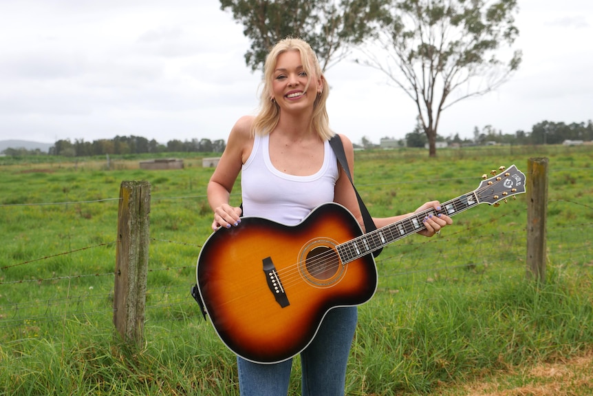 A woman with blonde short hair wearing a white singlet and jeans with a guitar strapped around her standing in a paddock