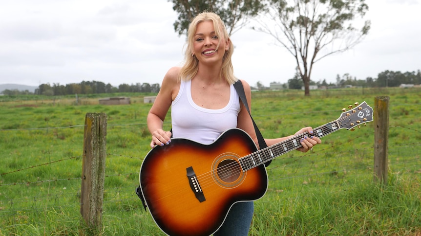 A woman with blonde short hair wearing a white singlet and jeans with a guitar strapped around her standing in a paddock