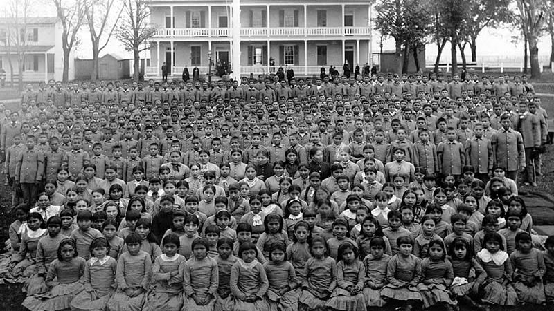 Indigenous children sit in a large crowd in front of a school building.