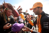 F1 driver Daniel Ricciardo leans over a barrier to sign a fan's purple cap as someone else holds a shoe to be autographed. 