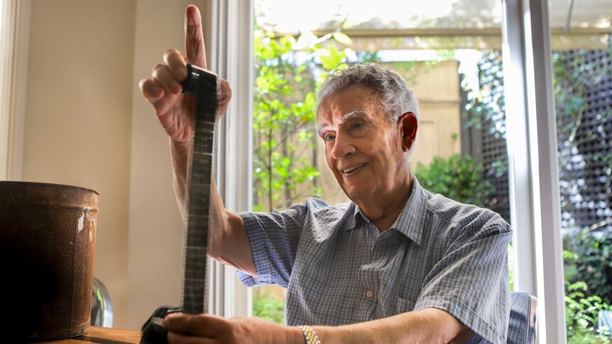 Con Petalas, an older Greek man with grey hair, holds a roll of film sitting at a wooden table with a garden in the background