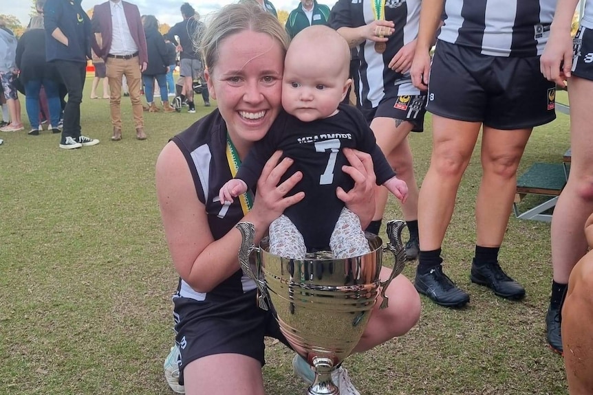 A young woman squats down with a large trophy and holding a small baby.