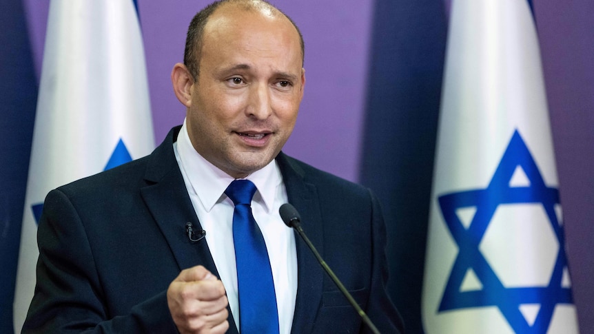 Naftali Bennett speaks behind a lectern with Israeli flags in the background. 