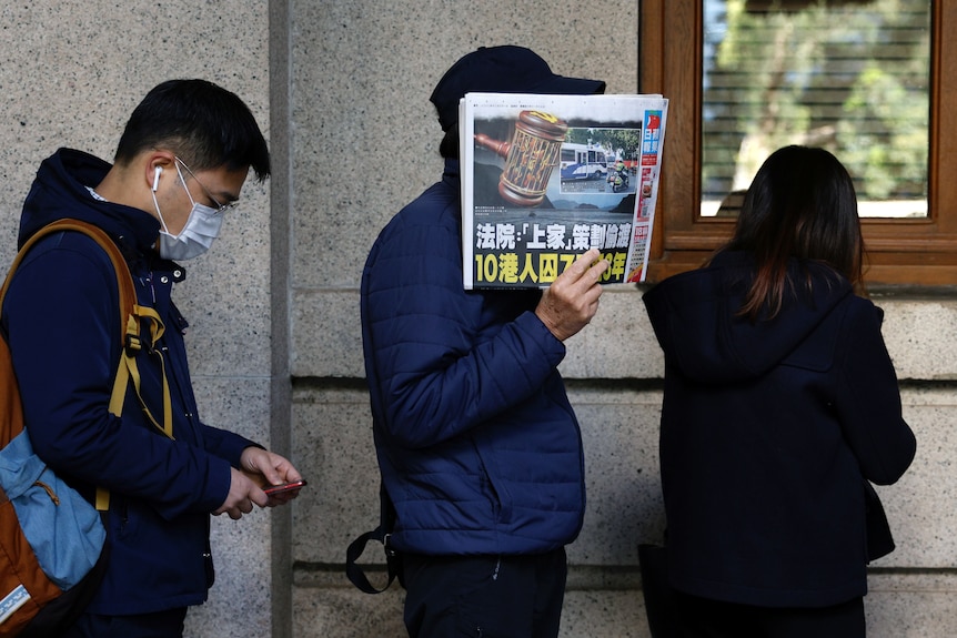 people stand in a queue as someone in the middle holds up a copy of a newspaper