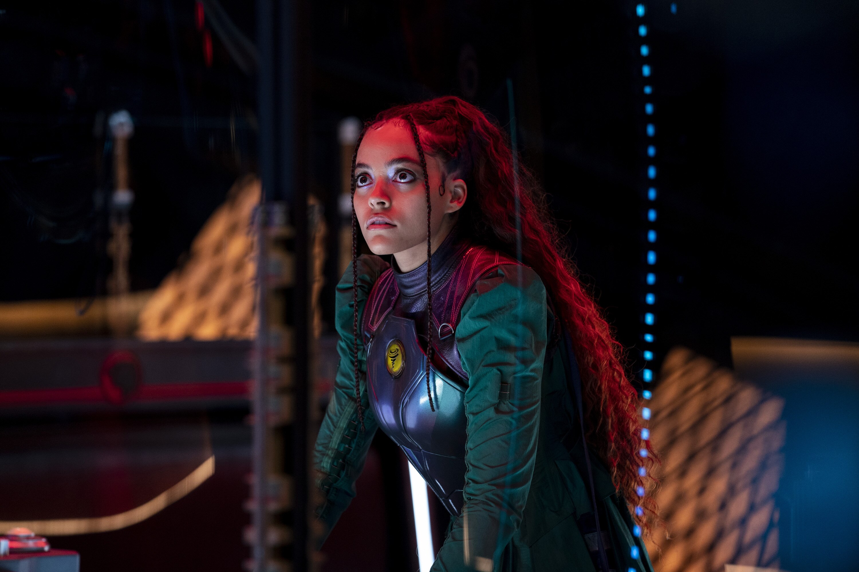 A young Black woman, with long hair, has a determined expression. She is wearing a top with a breastplate, with green sleeves. 