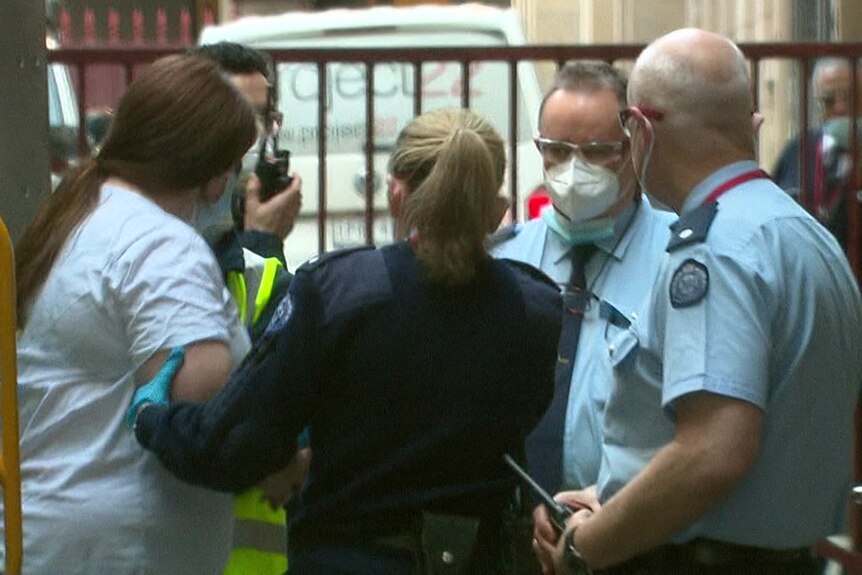 An overweight woman wearing a white t-shirt and a face mask is led away by police.