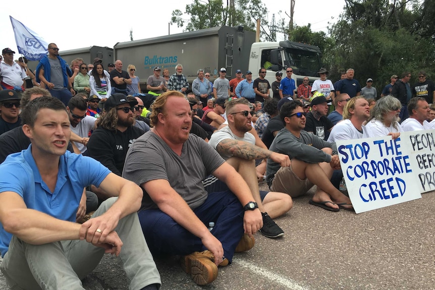 a group of men and woman, some with placards, sitting on road an chanting slogans