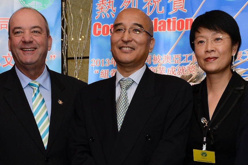 Two men in suits and one well dressed lady pose in front of Chinese-language banners.