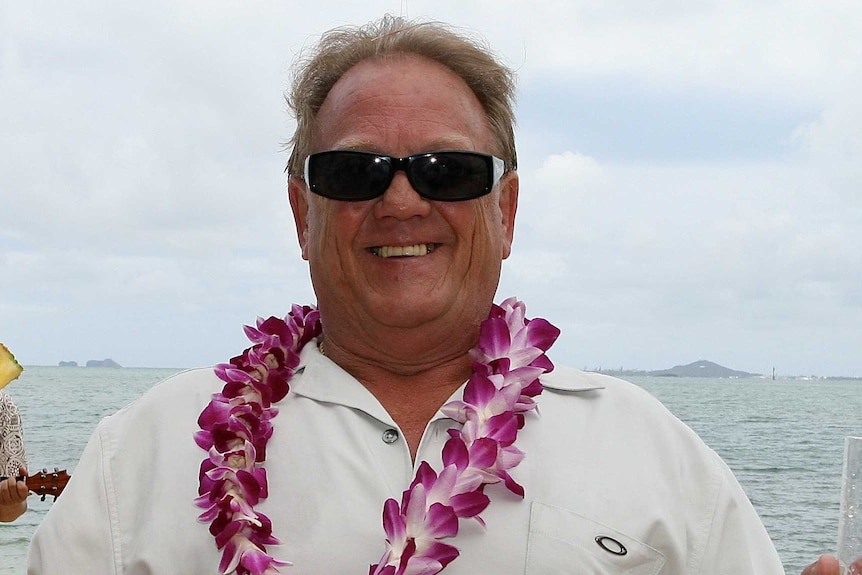 A close up of Peter Davis wearing a white shirt with pink flowers around his neck.