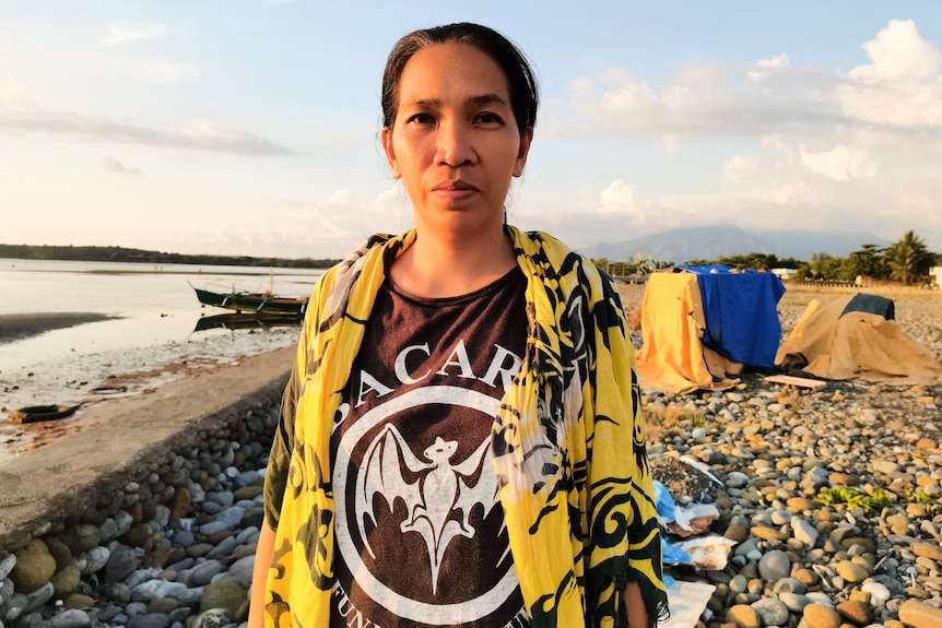 A woman with dark hair and eyes wears a tshirt and shirt while standing near the ocean.