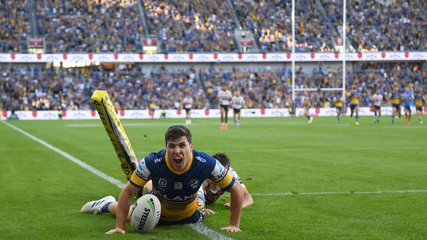 Mitchell Moses screams in delight as he slides over the try line with both hands on the grass, a stadium in the background.