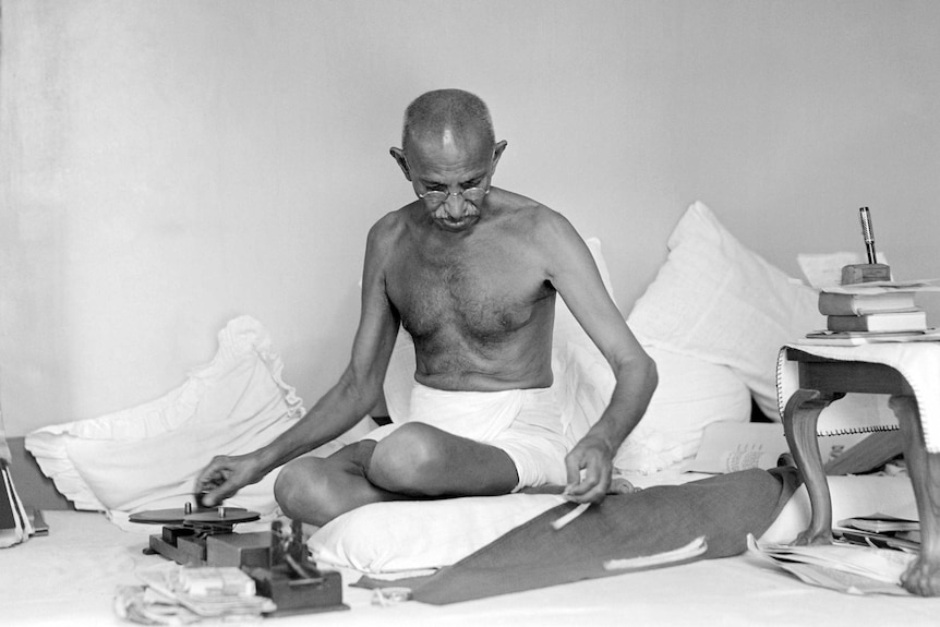 Mahatma Gandhi sits cross legged on the floor surrounded by pillows, cloth and a few books on a small table.