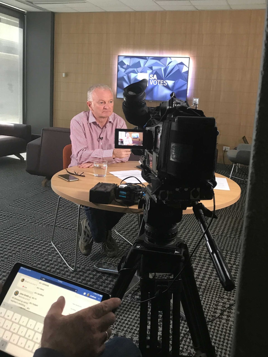 Antony Green sitting in front of TV camera with SA Votes screen in background and facebook on ipad in foreground.