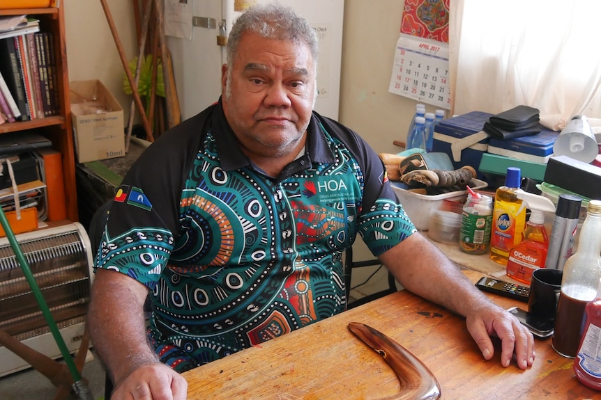 A man, looking serious, stares into the camera from his kitchen table, which has a boomerang on it.