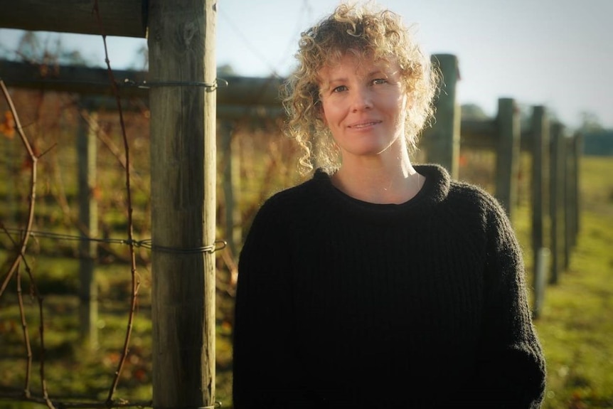 A woman with blonde curly hair in a black jumper stands by a post holding vines in a vineyard.