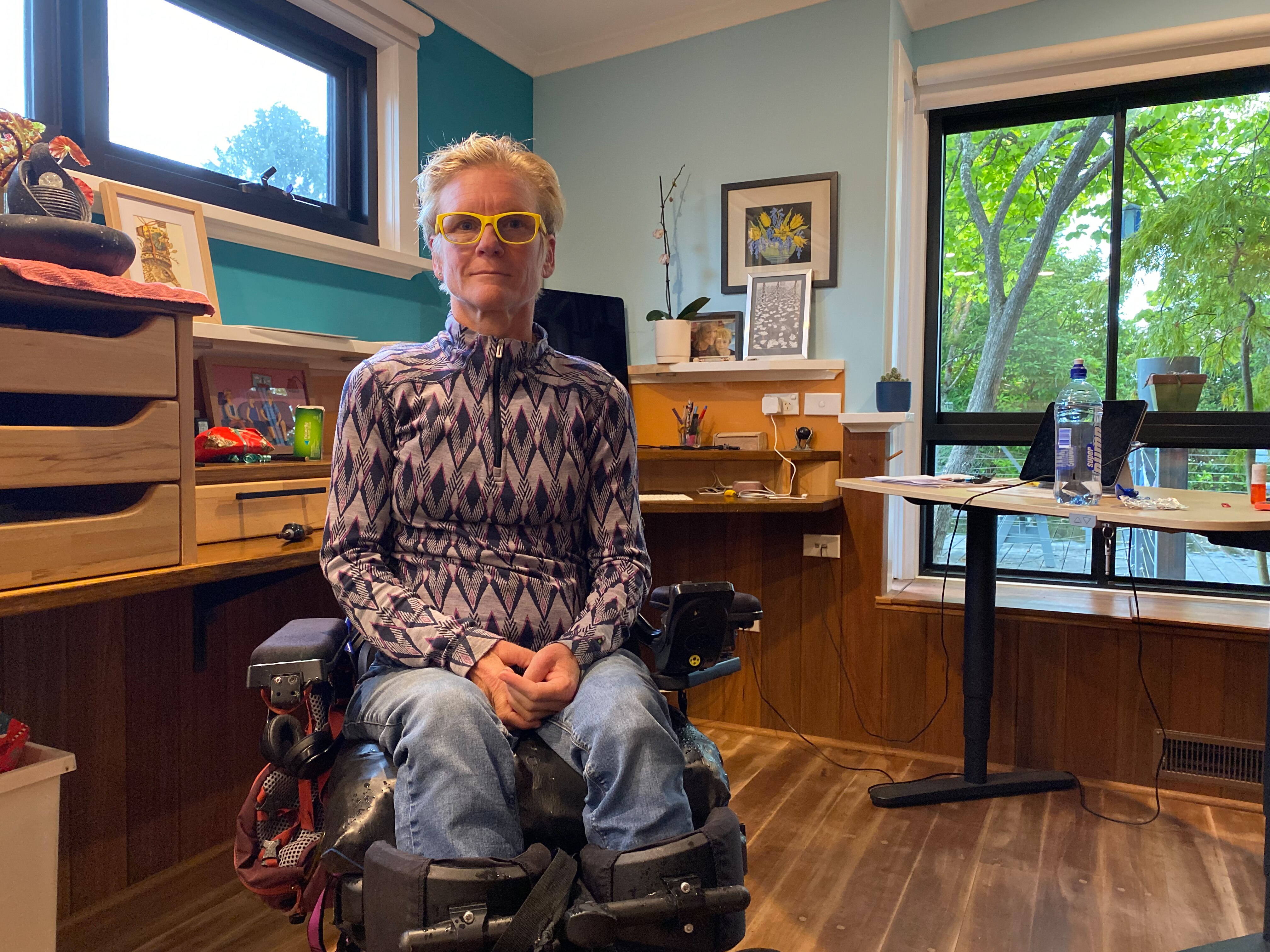 A woman wearing a geometric pattern top and yellow glasses sits in her wheelchair in a home study