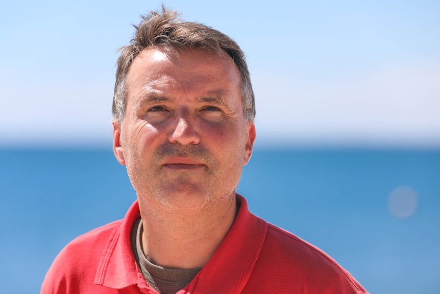 A middle aged man in a red shirt has a neutral expression standing on sand in front of the ocean.
