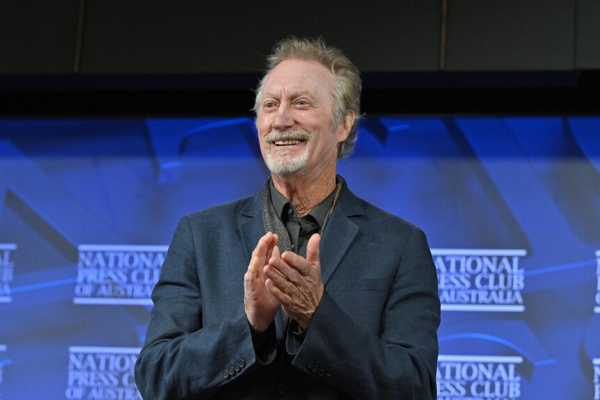 Bryan Brown smiling and clapping with a blue background, wearing a dark shirt, National Press Club of Australia written behind
