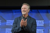 Bryan Brown smiling and clapping with a blue background, wearing a dark shirt, National Press Club of Australia written behind