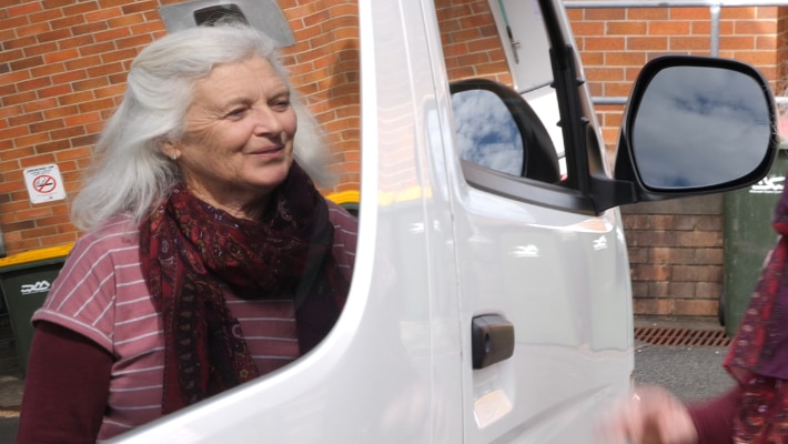 A woman with shoulder-length grey hair and a pink top, reflected in the window of a white van