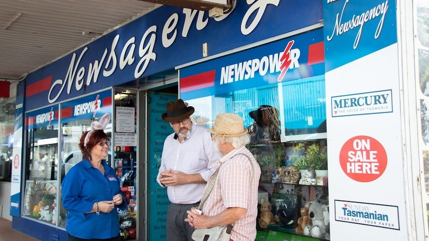 Man standing outside a newsagency talking to a woman and a man