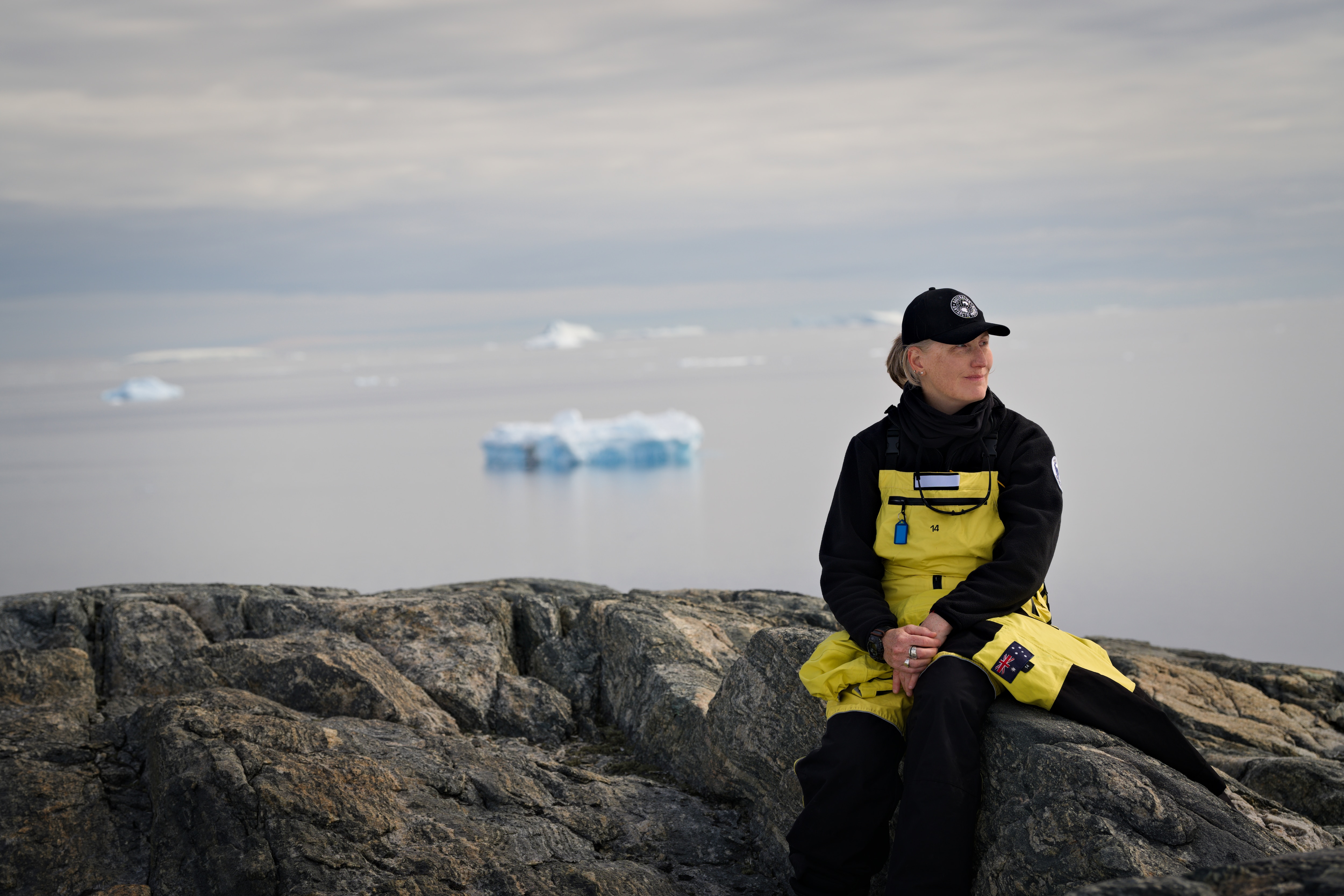 Annalise Rees senta em uma pedra e olha para o lado. Ao fundo você pode ver água e gelo marinho.