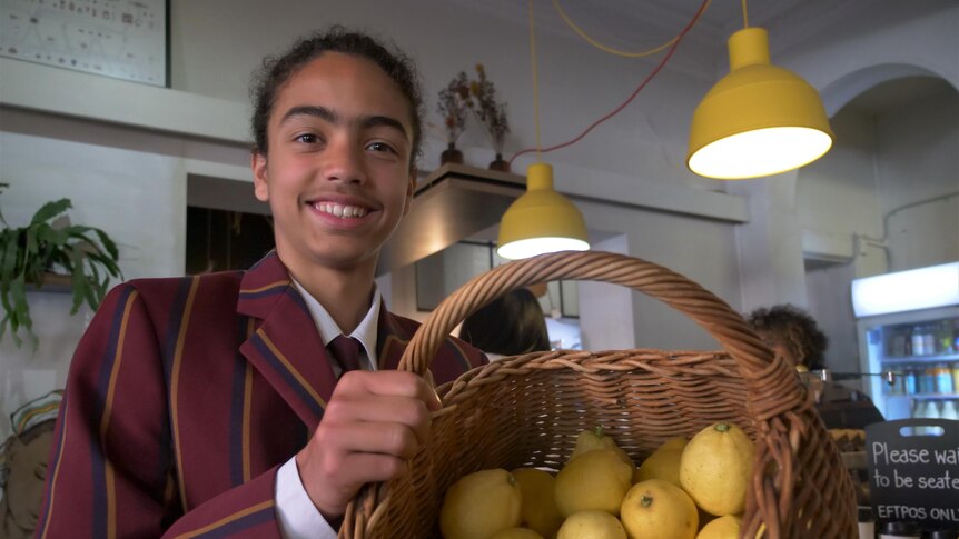 A teenaged boy holding a big basket full of lemons while standing in front of a cafe