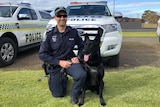 A man in police uniform kneels next to a black dog with police cars behind them