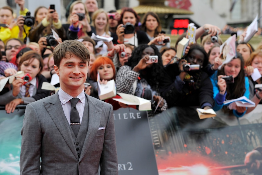 Daniel smiles at the film premiere in a grey suit as fans stand behind a barrier taking photos and holding Harry Potter books