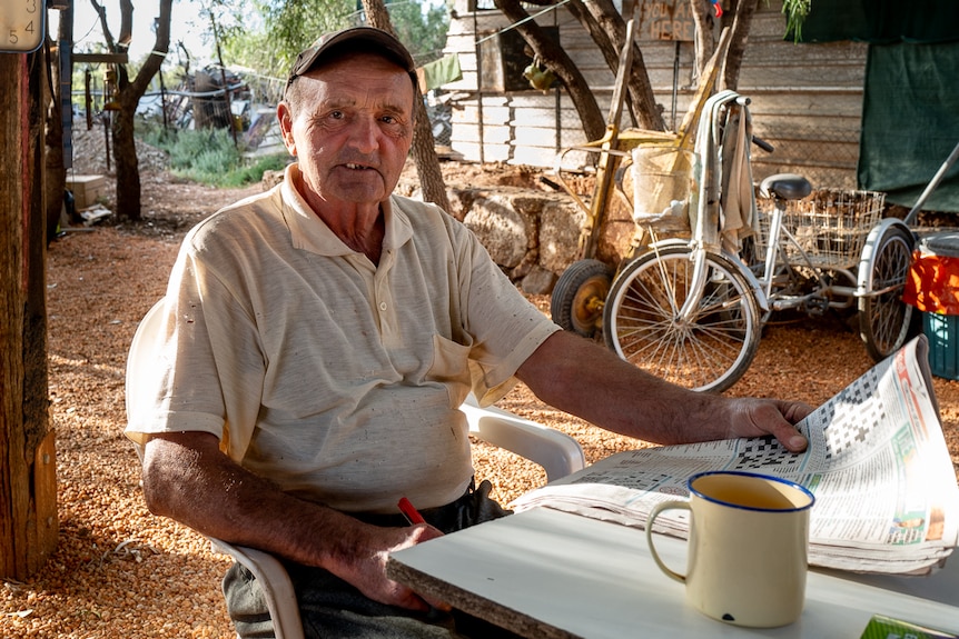 A man sits in a chair at his property with a newspaper and cup of coffee