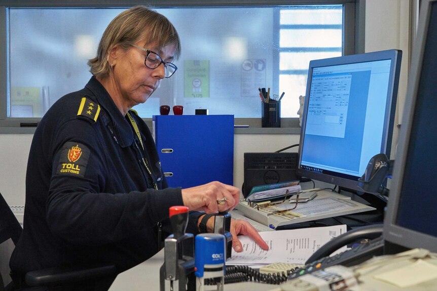 A blond woman in Norwegian 'toll customs' uniform stamps a paper form at her desk looking out into icy-blue light.